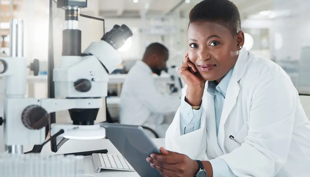 woman in white lab coat sitting at a table looking at a tablet