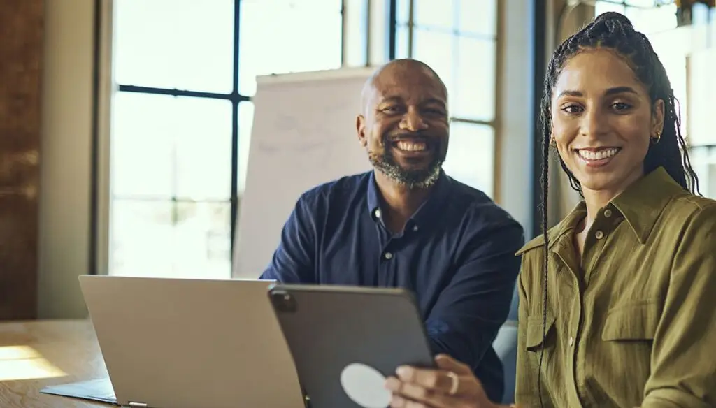 two smiling people sitting at a desk with a laptop computers