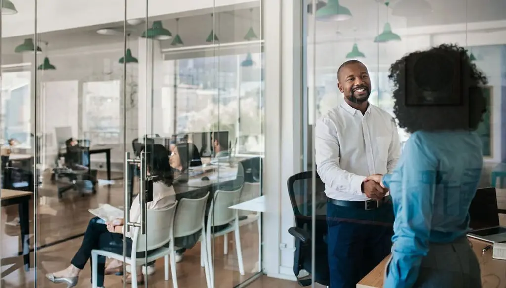 two people shaking hands with a meeting behind them
